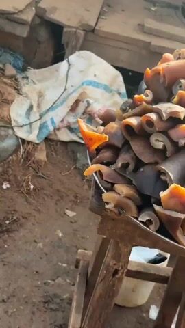 A video captures a marketplace scene, likely in a developing country, with a focus on food items and market stalls. The foreground features a close-up of a large metal bowl overflowing with various pieces of cooked pig skin, commonly known as "pomo" in some African cuisines. The pig skin is a range of light brown to deep amber colors, indicating different stages of preparation or cooking.

The middle ground reveals the general environment of the market. Wooden structures, presumably stalls or vendor booths, dominate the scene. Some are made of rough-hewn planks, while others are covered with tarpaulins or large sacks for shelter. A person, possibly a vendor, is partially visible behind a stall, tending to leafy green vegetables. The vegetables are piled high, suggesting fresh produce for sale. The overall impression of the market is one of activity and commerce, though the structures appear rudimentary and somewhat aged.

The background shows more of the market's architecture, with additional wooden stalls and coverings. There's a sense of depth and the presence of other market activities, though they are less defined. The lighting suggests it might be daytime, with sunlight filtering through the structures, creating some areas of shadow and brightness. The atmosphere appears to be that of an outdoor market, with the textures of wood, fabric, and produce being prominent.