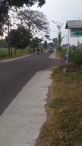 A black SUV is seen driving down a road, followed by a white motorcycle carrying one person. The motorcycle is on the right side of the road and the SUV is in the left lane. The road is paved with asphalt and there is a sidewalk on the right side. The area is rural, with trees and fields on either side of the road. The sky is overcast. There is a sign on the right side of the road that says "DUSUN SIDE".