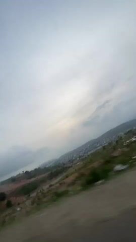 The video shows a landscape from a moving vehicle. The sky is overcast with white and grey clouds. A body of water is visible in the distance, partially obscured by clouds. A hillside covered in reddish-brown earth and sparse green vegetation slopes down towards the water. In the far distance, there is a densely populated area that extends up the hills. The road is visible in the foreground as the vehicle travels along it. There is a utility pole that goes by in the view.