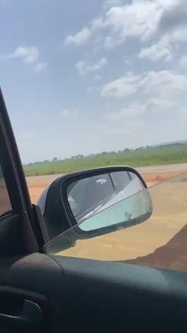 The video shows the view from inside a car traveling on a highway. The landscape visible through the car windows and side mirrors consists of flat, grassy fields under a partly cloudy sky. There is a cracked windshield in the car. Occasional vehicles can be seen in the distance on the highway. The interior of the car is also visible, including the dashboard, side door, and air vents.