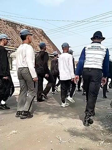 The video shows a group of young men in matching grey caps and athletic wear marching in formation on a paved road. They are all facing the same direction and are moving in sync, with their arms swinging. The men are dressed in various colored shirts, mostly white and black, with some wearing grey or blue tops. They are wearing casual pants, some with stripes down the side, and athletic shoes. Behind the marching group, a house with a tiled roof and an ornate fence is visible, along with utility wires and poles overhead. The ground appears to be a residential street. The overall atmosphere suggests a practice drill or a parade.