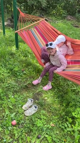 A child in a pink hoodie, patterned leggings, and a white hat with cat ears is lying in an orange and striped hammock. The child is smiling and looking at the camera. There is another child in the hammock, but only their head is visible. Two pairs of shoes are on the ground beneath the hammock. The hammock is suspended between two trees in a grassy, wooded area.