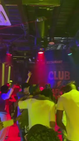 The video depicts a crowded nightclub with people dancing under flashing red, green, and yellow lights. The ceiling is visible with lighting equipment and ductwork. A sign with the words "CLUB" and "RESTAURANT" can be seen in the background. People are wearing casual clothing and enjoying themselves. The atmosphere is lively and energetic.