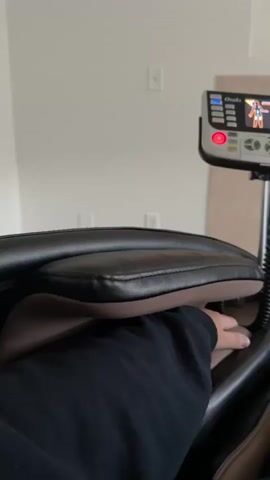 The video shows a person's legs and feet in a massage chair. The chair has a control panel with a screen displaying an animated figure and massage intensity levels. The person is wearing black pants and grey socks. The background appears to be a living room with a fireplace. The video is shot from a first-person perspective.