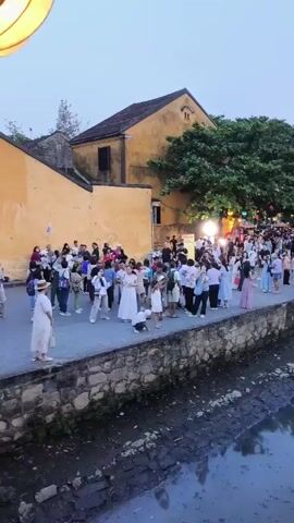 The video shows a crowded riverside scene in a town, likely in Asia. A concrete embankment lines a narrow waterway.  Numerous people walk along the embankment, with some appearing to pose for photos. A small bridge is visible in the distance, also filled with people. Buildings with Asian architectural styles are visible in the background, some adorned with lights. The sky is overcast and light.
