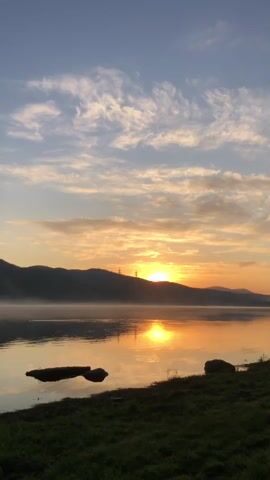 The video shows a sunrise over a calm lake. The sun is low on the horizon, casting a golden glow on the water and the sky. The sky is a mix of blue and orange, with wispy clouds. A line of hills forms the background, shrouded in mist. In the foreground, there is a grassy bank with a few rocks. The overall impression is one of peace and tranquility.