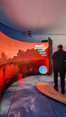 Here's a detailed description of the video:
The video shows a man standing on a surfboard in what appears to be an interactive surfing simulator. He is facing a large, curved screen displaying a vibrant landscape with red and orange hues, resembling a sunset over a body of water with mountains in the background. The man is dressed in a black button-down shirt and black pants. He is standing still on the surfboard, which is positioned on a floor designed to look like water.

To the right of the screen is a display panel with a "Top Finish Times" leaderboard. On the screen, instructions appear: "Right trigger to activate throttle" and "Lean left and right to influence direction," accompanied by illustrative arrows.

The room has a curved ceiling with recessed lighting, and there's a camera mounted above the screen. The overall environment is brightly lit.