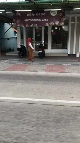 A woman wearing a red hijab and a tan dress walks in front of a storefront. The storefront has a glass door and windows. A sign above the door advertises a promotion: "Beli Rp. 100.000 GRATIS SQUARE CAKE". Two motorcycles are parked in front of the storefront. The scene appears to be on a street with a paved road in the foreground.