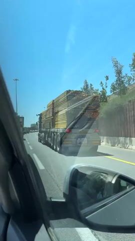 The video, shot from a moving vehicle, shows a large flatbed truck carrying a tall stack of wood or lumber moving along a highway. The lumber is banded together in stacks and appears to be yellowish in color. A concrete mixer truck passes on the right. The sky is clear and blue. The scene gives the impression of being somewhere sunny, possibly a highway with construction or development nearby.