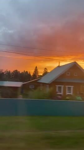 The video shows a view from a moving vehicle during sunset. The sky is filled with vibrant orange and pink clouds. In the foreground, there are glimpses of houses, fences, trees, and greenery. The motion blur indicates the perspective of someone traveling.