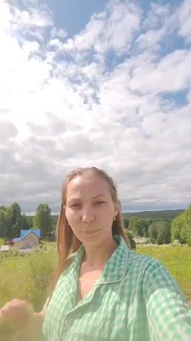 A woman with long blonde hair, wearing a green and white checkered shirt, smiles and waves at the camera. She is standing outdoors in a grassy field with trees in the background and a blue sky with white clouds overhead. There are a few small buildings and a dirt road visible in the distance. The sun is shining brightly.