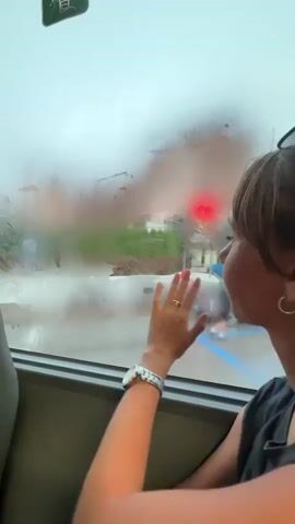 The video shows a person inside a vehicle looking out a condensation-covered window. The person's right hand is on the window, touching the condensation. Outside, through the fogged-up window, a blurry outdoor scene is visible, with buildings and what appears to be a street or parking lot. It is raining or humid, causing the condensation on the window.