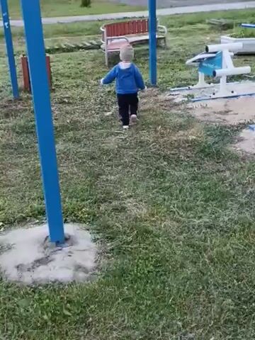 This video, recorded in Smolino, Russia, captures a young child in a blue jacket and hat walking across a grassy outdoor area. The child is walking towards the right side of the frame. In the background, there is a house with an orange facade and white window frames, a wooden fence, and several trees. A metal structure with a transparent roof, possibly a gazebo or a covered play area, is prominent in the foreground, with blue metal poles framing the scene. To the right, there are some outdoor exercise equipment structures, one white and light blue, and a bench. The scene appears to be set in the late afternoon or early evening, with overcast or cloudy weather. The overall atmosphere is calm and domestic, suggesting a residential area or a park.