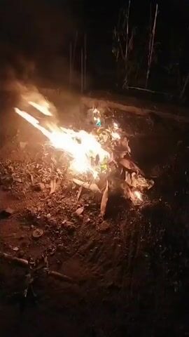 A small campfire burns in the dark, with flames of yellow and orange consuming dry leaves and twigs. The fire is surrounded by dark, uneven ground, possibly dirt or ash. In the background, the outlines of trees or other vegetation are visible against the blackness of the night sky. The overall scene suggests a rustic or outdoor setting at night.