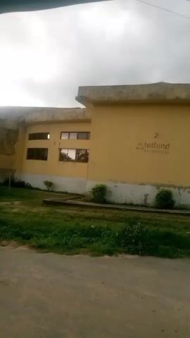 The video pans from left to right, showing a yellow building with a curved section and windows. The building has the word "tetfund" written in grey on the right side. A person wearing a black top and brown skirt walks across the frame from left to right in front of the building. The building is surrounded by green grass and a paved path. The sky is overcast.