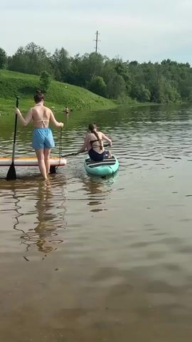 Two young women are preparing to paddleboard on a calm lake. One stands on a yellow paddleboard close to the shore, holding a paddle and what appears to be a coiled leash. She is wearing a light-colored bikini top and blue shorts. The other woman is already seated on a blue-green paddleboard, using her paddle to maneuver away from the shore. She is wearing a dark swimsuit. The lake is surrounded by green trees and grassy banks. In the background, a few people are visible on the shore, and there's a power line tower in the distance. The water is reflecting the sky and the surrounding greenery. Part of a person wearing a red cap is visible in the foreground.