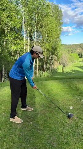 A golfer in a blue shirt, black pants, and a beige baseball cap is on a golf course. He tees off and hits the ball with his driver. The ball travels across the course. The golfer watches the ball, then looks back at the camera. The golf course is green and has trees, and it is a sunny day with a blue sky.