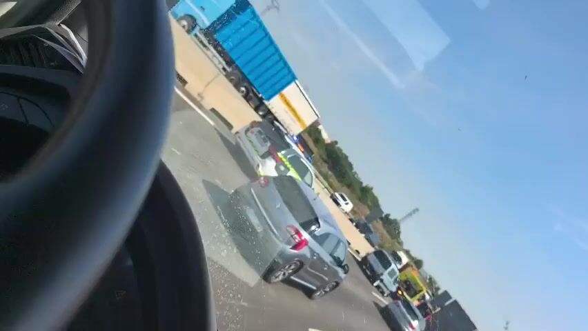 The video shows a traffic accident on a highway from the perspective of someone behind the wheel of a vehicle. Multiple cars and a police vehicle are stopped on the side of the road, and a tow truck is present. The weather appears to be sunny.  The video is shot from an interior perspective, showing part of the steering wheel.