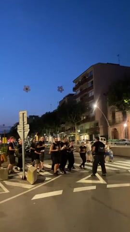A group of people in black shirts are gathered on a street corner at dusk in Arenys de Mar, Spain. They appear to be performing music, as many are holding instruments that resemble drums and drumsticks. Some individuals are actively striking their instruments, creating a rhythmic beat. The sky is a deep blue, indicating the time of day is early evening. In the background, there are buildings, trees, and streetlights, suggesting an urban setting. Two large, stylized floral decorations are strung across the street overhead. The people seem engaged in their performance, with some smiling and looking towards the activity. The overall atmosphere is one of a lively street performance or gathering. There is no visible text in the media file.