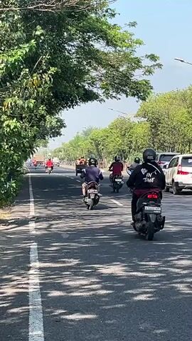 The media file captures a dynamic scene on a road in Ketapang, Indonesia, likely during daytime. A convoy of motorcycles, carrying both riders and passengers, moves along the asphalt. The motorcycles are diverse, with some appearing to be scooters and others more traditional models, many in various colors like red and black. The riders are predominantly wearing helmets, some with distinctive designs, and casual clothing.

The road is bordered by lush green trees on both sides, their leaves casting dappled shadows across the pavement, indicating a sunny day with ample sunlight. The sky is clear and bright, suggesting fair weather. In the background, glimpses of other vehicles, including cars and possibly more motorcycles further down the road, are visible, hinting at moderate traffic.

The perspective is from a moving vehicle, likely another motorcycle or a car, following the procession. The overall mood appears to be one of casual travel or perhaps a group outing. The subjects are focused on their journey, with no overt expressions of emotion readily apparent.

The scene is clearly outdoors, on a paved road with a white line demarcating the lanes. The presence of trees and the style of vehicles suggest a tropical or sub-tropical environment. No specific text is legible on the vehicles or clothing. The activity is transportation, with multiple individuals traveling together on their motorcycles.