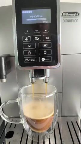 A close-up, eye-level shot shows an automatic coffee machine, a DeLonghi Dinamica, brewing coffee into a double-walled glass mug. The machine's display screen reads "My Kaffee" and "STOP". Two streams of dark brown coffee are pouring from the spouts into the mug, which is already partially filled with coffee and topped with a layer of light brown foam. The coffee machine is silver and black, with various buttons and a digital display. The mug is transparent and has a handle. The coffee machine sits on a silver drip tray. The background is blurred and appears to be a kitchen counter.