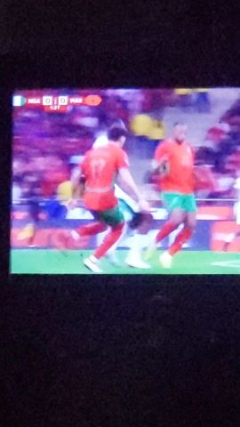 A vertical video shows a soccer game being played on a green field. On the left of the screen is the left third of the television, including the scoreboard. The stadium has a crowd in the background. Players run across the field.
