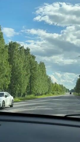 The video captures a forward-facing view from inside a vehicle, likely through its windshield, as it drives along a straight asphalt road in Imatra, Finland. The road is clear, with two visible lanes, and stretches into the distant horizon.

On the left side of the road, a continuous line of tall, lush green trees, likely birches, forms a dense natural barrier. On the right side, a similar line of trees is visible, along with glimpses of streetlights and possibly power lines, suggesting some roadside development. A dark-colored vehicle is discernible in the far distance, traveling in the same direction.

The sky above is a mix of bright blue patches and large, fluffy white and grey clouds, indicating a partly cloudy day. The sunlight is strong, casting subtle shadows on the road surface. The overall impression is that of a pleasant daytime drive. The ambient sound consists of a steady vehicle hum and road noise. No people or visible text are present within the scene.