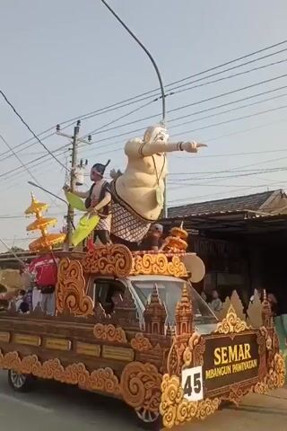 A parade float moves forward under a clear sky. On the float, two people are positioned on a raised platform decorated with ornate gold carvings. The person on the left is a male dressed in traditional clothing, including a headdress, a patterned sash, and a yellow cloth draped around his neck. He is holding a staff and gesturing with his other hand. To his right, a large, light-skinned effigy of a person with exaggerated features and a prominent belly is seated. This effigy is also pointing with its right arm. The float itself is a wooden structure with decorative carvings on its sides, and a banner with Indonesian text is visible. In the background, houses and utility poles suggest an outdoor, possibly urban or semi-urban, setting. A few spectators are visible in the foreground, and a small Indonesian flag is attached to a pole on the float.