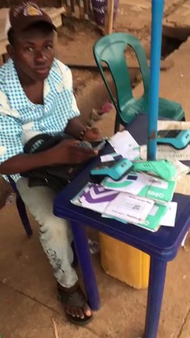 The video shows a man seated behind a small, blue plastic table, working on his phone. The table is cluttered with receipts, what appear to be point-of-sale (POS) devices, and some cash. The man is wearing a blue and white checkered shirt, jeans, and a brown baseball cap with "CHAMPS" and the Lakers logo on it. He has a black backpack resting on his lap. A green plastic chair is nearby. The setting appears to be an outdoor market area, with a dirt and concrete surface and some basic shelter overhead.
