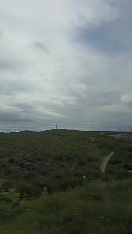 A video captures a cloudy sky over a grassy, hilly landscape. The camera appears to be in motion, possibly from a vehicle, as the grassy foreground moves in a blurred manner. In the mid-ground, there are several large rocks and some sparse vegetation. In the distance, there are at least two utility poles visible. The overall impression is of a rural, possibly coastal, environment under an overcast sky. The video is of short duration, approximately four seconds.