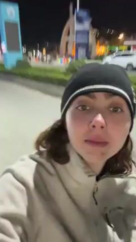 A young woman, wearing a black beanie with a white stripe, is filming a selfie video at night. She is standing on a street with cars driving by. Streetlights and building lights illuminate the scene. There's a traffic circle in the distance. The woman's gaze shifts around the scene.