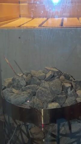 The video shows a close-up view of a sauna heater filled with rocks. A red glowing element is visible within the rocks, indicating the heater is on. A thin, red stick, possibly an incense stick, is placed among the rocks. In the background, a wooden bench is visible, along with a bright light source reflecting off a glass or mirror surface. A hand reaches into the frame from the right and places a dark-colored cup on the bench before retreating. The overall environment appears to be a dimly lit sauna.