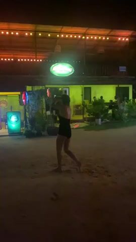 A medium shot depicts a person wearing a black top and shorts standing barefoot on a sandy surface at night. They are facing away from the camera and appear to be performing a poi dance with glowing objects in their hands. Behind them is a building with a green neon sign that reads "Chang". The building is lit with yellow light, and a string of orange lights hangs above. Several people can be seen sitting inside the building. The person is mostly stationary, moving their arms to manipulate the poi. Another person walks by in the right side of the frame.