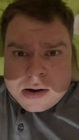 The video shows a close-up of a man lying down. He is wearing a gray shirt with buttons. The background is a green pillow. The man's face fills most of the frame, and he appears to be speaking or reacting to something. His mouth is open, and his eyes are wide. He occasionally touches his head.