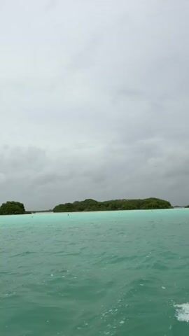 The video shows a view from a boat moving through turquoise water. In the distance, there are several small islands covered in green vegetation. The sky is overcast with gray clouds, and there are numerous small dark specks, likely birds, visible in the sky. The boat is creating a wake behind it, visible in the foreground. The overall impression is a serene but slightly somber coastal landscape under a cloudy sky.