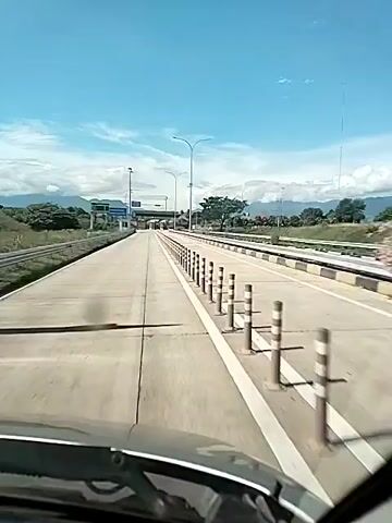 The video shows a view from inside a car driving on a toll road. The sky is blue and clear, with some wispy clouds in the distance. The road is straight and wide, with white lines marking the lanes.  There is a line of black and white bollards separating the lanes. Streetlights are on either side of the road. In the distance, there is a toll booth and some trees. The environment appears to be a rural or suburban area, with mountains visible in the distance.