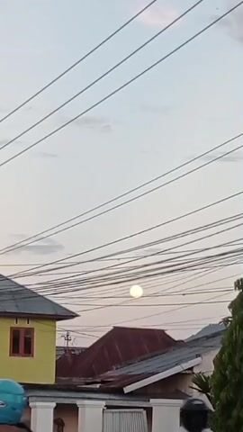 The video shows a bright, full moon visible in the sky above a residential area. Several power lines are strung across the frame, obscuring parts of the sky and the moon. In the foreground, parts of houses with yellow and red roofs are visible. The sky is a pale, hazy color, suggesting it might be dusk or dawn. A few people are partially visible at the bottom of the frame, some on motorcycles, suggesting activity in the street below. The overall scene is a snapshot of an urban or suburban environment at a time when the moon is clearly visible.