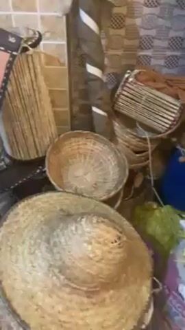The camera pans around a room showcasing an assortment of handmade musical instruments, mostly drums.  The drums are painted blue with yellow bands around them and appear to be made from recycled plastic barrels.  There are also woven baskets of various sizes, a woven cylinder, and what appears to be a woven fan, along with assorted fabrics.  The room has a tiled wall.