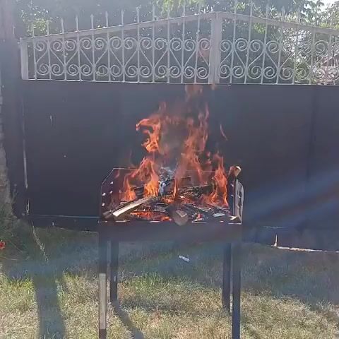 A campfire is burning in a portable metal grill. The grill is black and has a rectangular shape with legs. The fire is made of wood logs and is producing a lot of flames and smoke. The grill is situated on a grassy area in front of a black fence with a decorative white metal gate. The fence and gate appear to be part of a yard or outdoor space. The sun is shining, casting shadows on the ground. The overall scene suggests a casual outdoor setting, possibly for a barbecue or gathering.