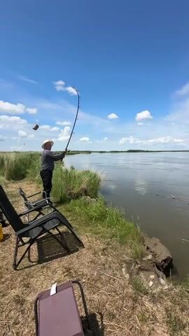 A person wearing a hat and gray shirt is fishing from the bank of a wide river under a blue sky with scattered white clouds. Two black folding chairs are positioned near the person, and a brown folding stool is in the foreground. The riverbank is covered in green grass and dry earth. The water is calm, reflecting the sky. In the distance, a treeline marks the opposite bank of the river. A small dark object is visible in the sky, possibly a bird or insect.