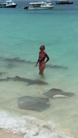 A woman stands in the clear, shallow turquoise water of a tropical beach, surrounded by several large stingrays. She wears a long-sleeved, high-cut swimsuit with an intricate pattern and sunglasses. With arms extended, she appears to be interacting with the rays as they swim around her legs in the gentle waves. The beach has light sand, and in the background, several boats are anchored in the distance. The water is clear enough to see the rays moving along the sandy bottom.