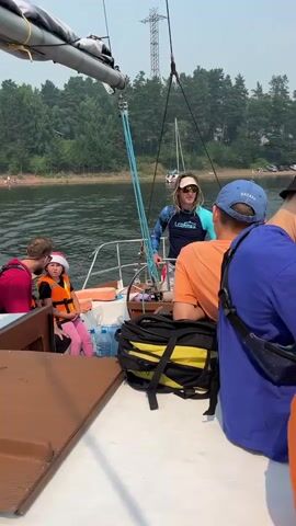The video shows a group of people on a sailboat. In the foreground, a man wearing a dark t-shirt is leaning on a winch. To his right, a man in a red shirt is looking back, speaking to a young girl wearing a pink hat and an orange life vest. The girl is sitting next to a pile of blue water bottles and a black and yellow bag. Behind them, a person wearing a blue and black long-sleeved shirt and sunglasses is steering the boat. Another person in a blue baseball cap is visible further back. The boat is on a body of water, with a tree-lined shore in the background. A transmission tower is visible in the distance. The sky is hazy.