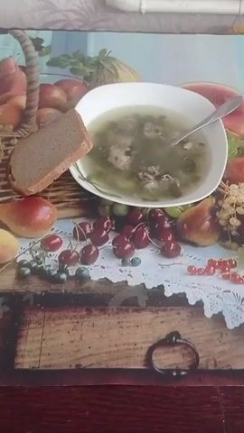 A white bowl of green soup, possibly sorrel soup or borscht, is centered on a tablecloth with a fruit and wooden board design. A metal spoon is submerged in the soup, resting on the rim of the bowl. A slice of dark bread is placed to the left of the bowl, standing upright. The tablecloth features various fruits, including cherries, blueberries, and red currants, clustered around the bowl, and larger fruits like apples, pears, and a watermelon slice are visible in the background. The scene appears to be an indoor setting with a light-colored wall and a pipe visible at the top. The video captures a still shot of the food.