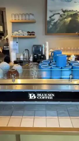 The video shows the counter of a coffee shop called "Blue Six Coffee". Behind the counter are two employees. One is wearing a black shirt and cap, while the other is wearing a white cap and a black shirt. A blue espresso machine is on the counter, with rows of blue mugs stacked on top of it. There is a shelf above the counter with glassware, cups, and other items. A landscape painting hangs on the wall.