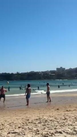 Three men are playing with a ball on a beach on a sunny day. They are all wearing swimming trunks. In the background, there are many people swimming in the water, and there are buildings and trees on a headland. The sky is blue and clear. The men are kicking and passing the ball to each other. The waves are gently lapping on the shore.