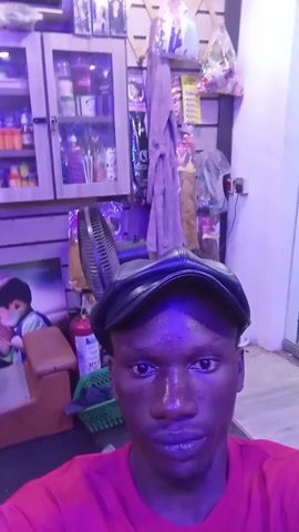A close-up video features a young, dark-skinned man wearing a red t-shirt and a black leather cap. He is indoors, standing in what appears to be a shop or small business, based on the merchandise displayed in a glass-fronted cabinet behind him. The shop is illuminated with a purple/blue light, which casts a noticeable tint over the scene and the man's skin. The man looks directly at the camera, and he briefly covers his mouth with his hand. In the background, various products and items are visible, along with a fan, a fire extinguisher, and a door with a "push out" sign.