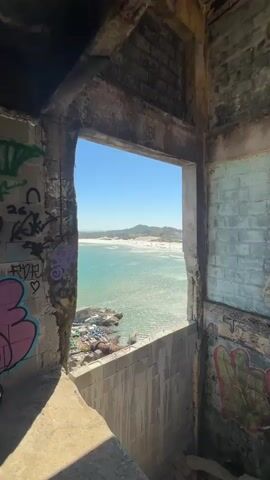 The video opens with a close-up shot of an opening in a ruined concrete structure. The opening frames a bright, sunny view of a coastline. The concrete structure appears to be heavily graffitied, with spray paint visible on the walls. Through the opening, a wide expanse of the ocean is visible, its surface shimmering in the sunlight. The water is a clear turquoise color. Beyond the ocean, a sandy beach stretches out, dotted with numerous people and umbrellas. In the background, rolling hills covered in vegetation rise, with a few buildings and structures visible on their slopes. The sky is a clear, vibrant blue. The camera then zooms out slightly, offering a wider perspective of the scene through the opening. The video then transitions to a wider shot, predominantly showing the beach and ocean. The camera pans slightly to the right, revealing more of the coastline and the distant hills. The beach is crowded with people, some sunbathing, others walking along the shore or swimming in the ocean. The water remains clear and inviting. The distant hills provide a scenic backdrop to the lively beach scene. The video concludes with a final shot of the beach and ocean, with the sun still shining brightly. The overall impression is of a beautiful, sunny day at a popular beach, viewed from the unusual perspective of a derelict building.