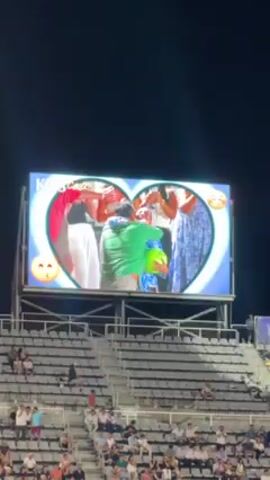 A large LED screen is displaying a heart-shaped image of a group of people, likely a family, embracing and celebrating. The image is enhanced with emojis, suggesting a joyous occasion. The screen is situated above stadium seating that is partially filled with spectators. The background is a dark night sky. The overall scene captures a moment of celebration being shared with a crowd.