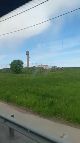 The video is shot from within a moving vehicle. Outside, the landscape is dominated by a field of green grass, dotted with yellow flowers, likely dandelions. Beyond the field, there's a fenced-off area containing a tall, concrete water tower. A small, leafy tree stands to the side of the tower.  A low building can be seen in the background, inside the fenced perimeter. The sky is blue with scattered white clouds. A roadside barrier is visible in the foreground, and a utility pole passes by briefly.
