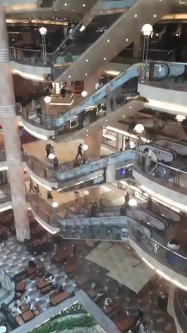 The video shows an interior view of a multi-level shopping mall. Escalators are visible connecting the different floors, with people moving on them. There are shops and storefronts along the curved balconies of each level. Some of the storefronts display advertisements for food, including burgers. People are seen walking around, shopping, and congregating in various areas. The mall appears to be well-lit with overhead lighting.